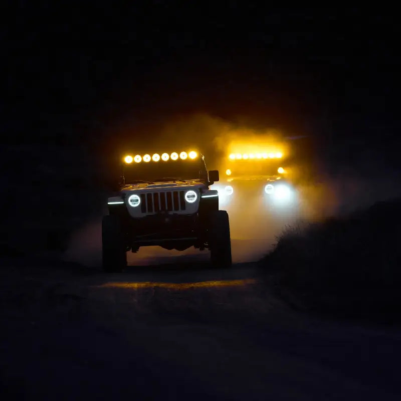 Two vehicles driving at night with the 50-inch Gravity Titan LED Light Bar emitting amber and white beams on a dusty trail.