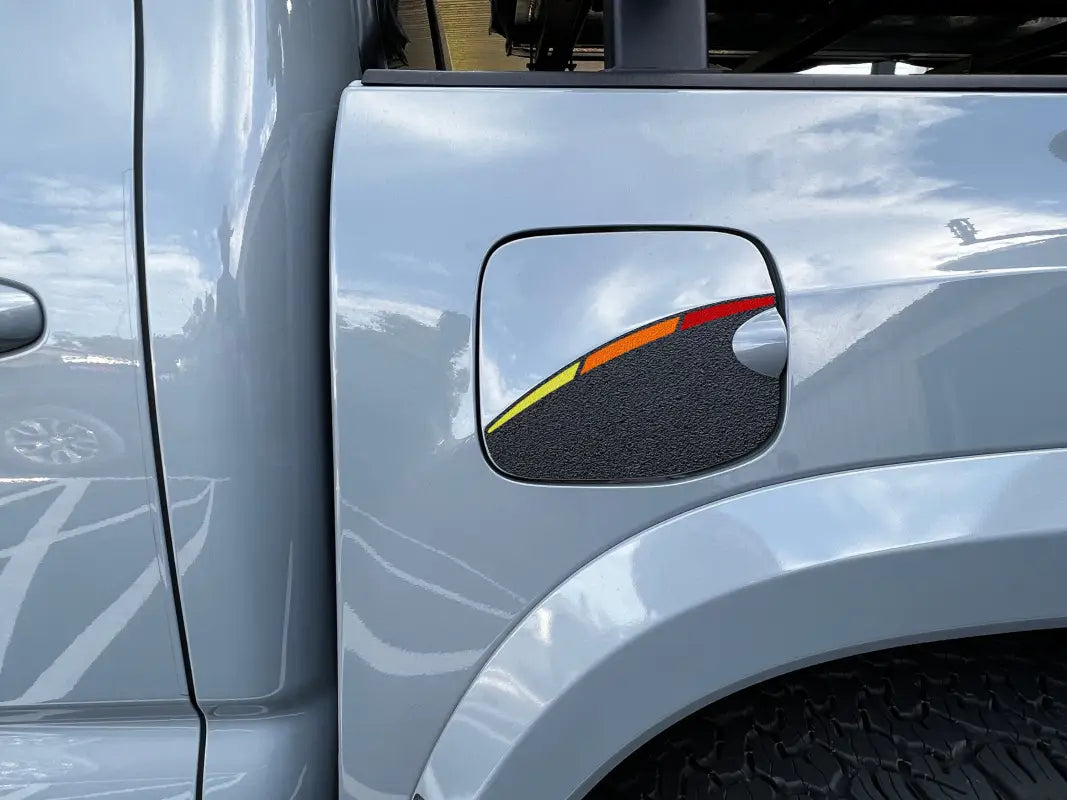 Gas lid protection overlay featuring black texture and tri-color stripe design in yellow, orange, and red, installed on a 2016-2023 Toyota Tacoma.
