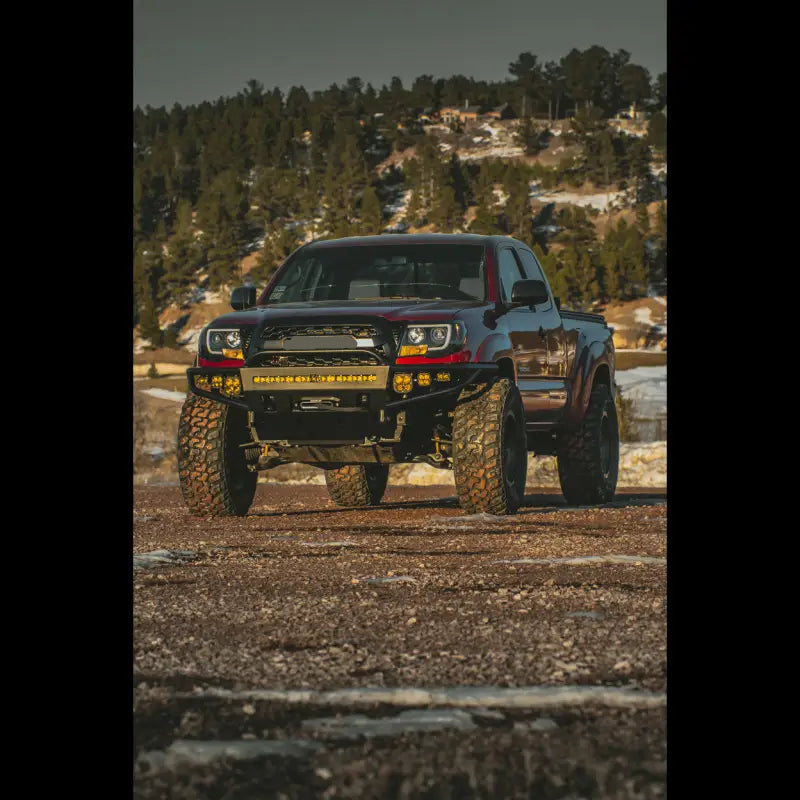 Red 2nd Gen Tacoma equipped with a hybrid front bumper, off-road tires, and winch-ready design, set against a forested background.