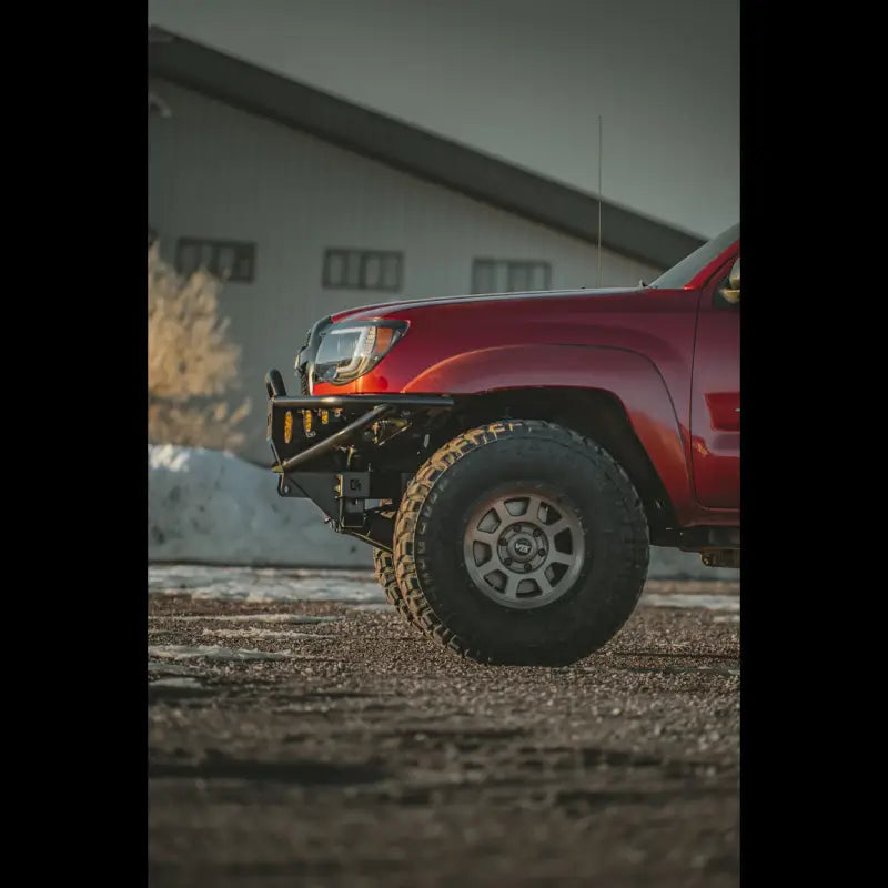 Side view of a red 2nd Gen Tacoma showcasing the hybrid front bumper and off-road tires, parked near a building.