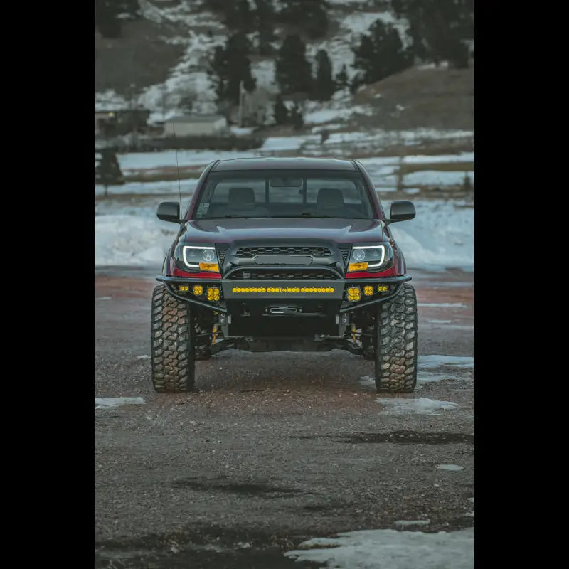 Front view of a 2nd Gen Tacoma with a hybrid front bumper, featuring off-road enhancements and integrated light bar, parked on snowy terrain.