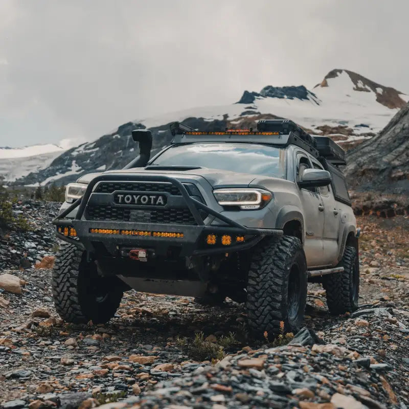 3rd Gen Tacoma equipped with a steel hybrid front bumper in an off-road mountain setting. Features winch compatibility and light bar support.
