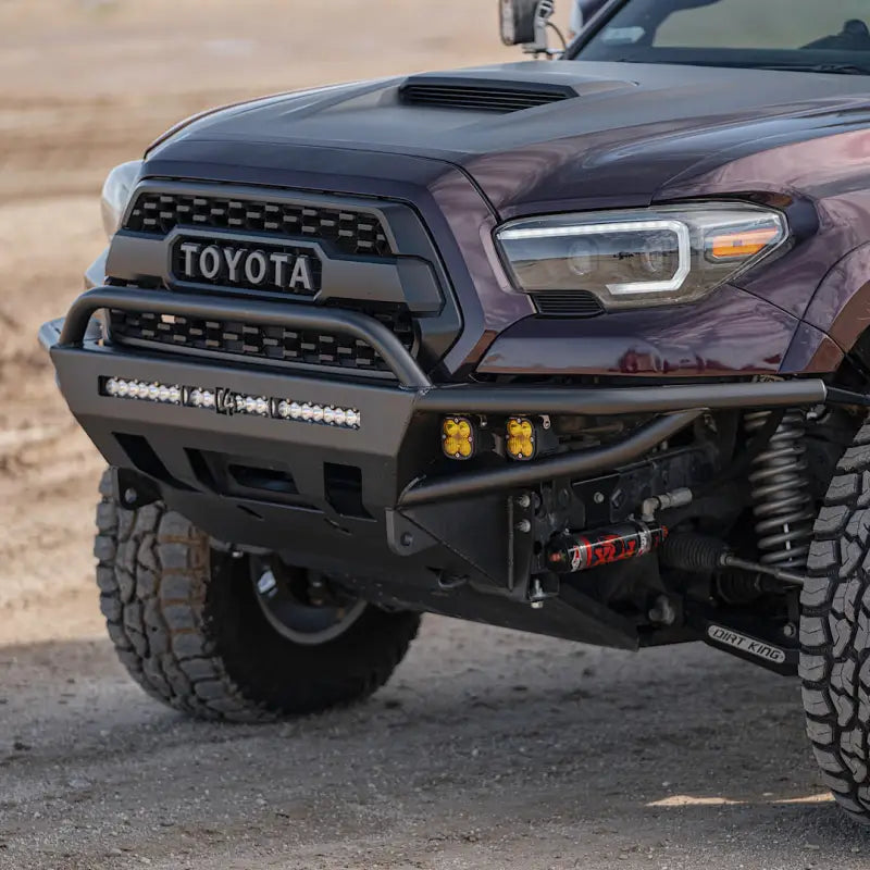 Close-up of a 3rd Gen Tacoma with a hybrid steel front bumper, featuring yellow fog lights and a winch mount in a rugged outdoor setting.
