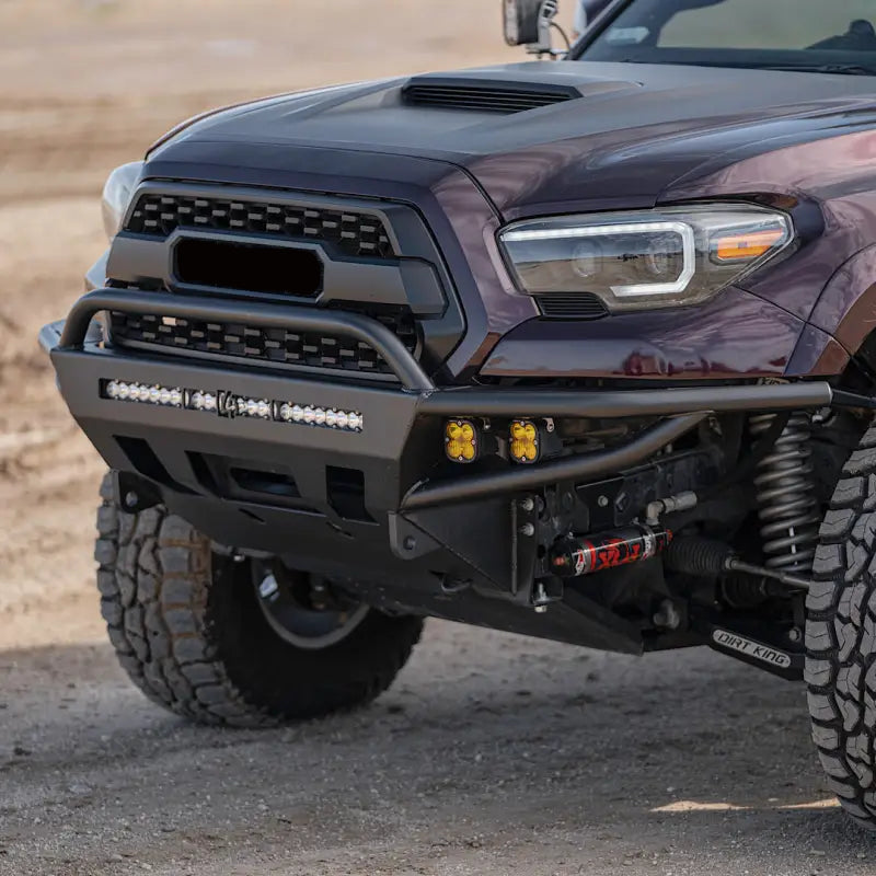 Close-up of a 3rd Gen Tacoma with a hybrid steel front bumper, featuring yellow fog lights and a winch mount in a rugged outdoor setting.