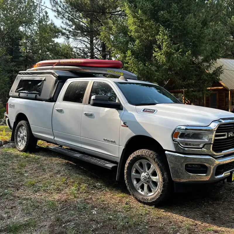 White truck with MR Full-Size Truck Topper carrying outdoor gear, parked near a cabin, highlighting its camping and adventure readiness.
