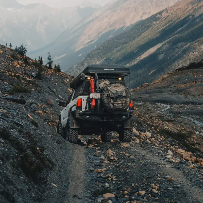 Tacoma Overland rear bumper on a 3rd Gen 2016+ Tacoma navigating rugged mountain terrain with camping gear and spare tire.