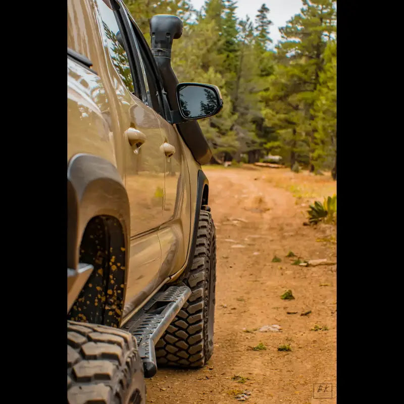 Side view of a 2016+ Tacoma with bolt-on rock sliders on a forest trail. Features durable design and hi-lift jack compatibility.