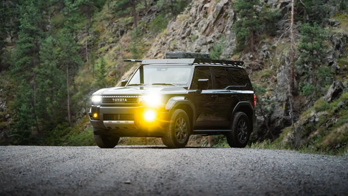 Front view of a black 2024 Toyota Land Cruiser with The Atlas roof rack, driving through a forested area with headlights on.