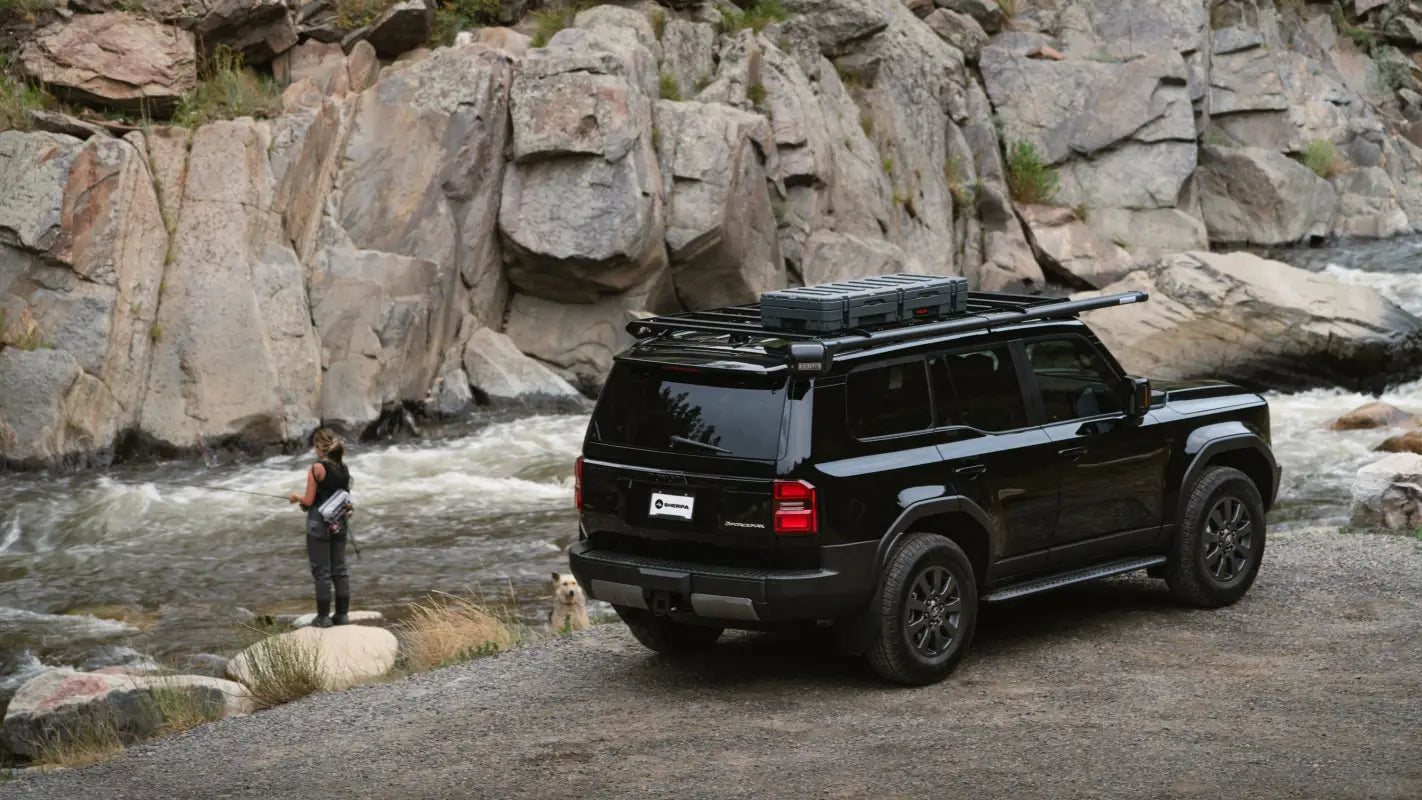 Rear view of a black 2024 Toyota Land Cruiser parked by a river, equipped with The Atlas roof rack and outdoor gear.