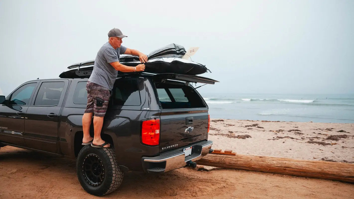 Man securing surfboards on the Crow’s Nest topper rack atop a black truck parked on a beach, showcasing its versatility for water sports gear.