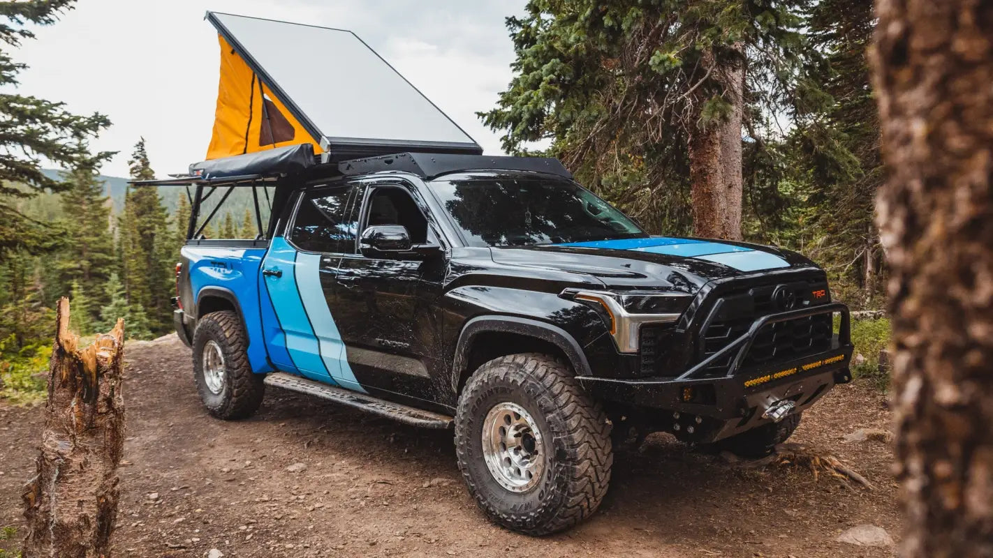 Toyota Tundra with The Cub Roof Rack and camper setup in a forest, featuring a pop-up tent and rugged off-road accessories.