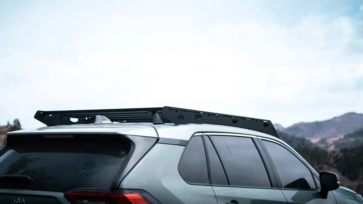 Rear side view of a gray RAV4 with The Snowmass roof rack, highlighting its full-length black aluminum construction in a mountain backdrop.