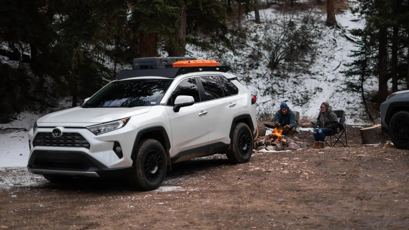 White RAV4 with The Snowmass roof rack parked in a snowy forest campsite, carrying recovery boards and a storage box.