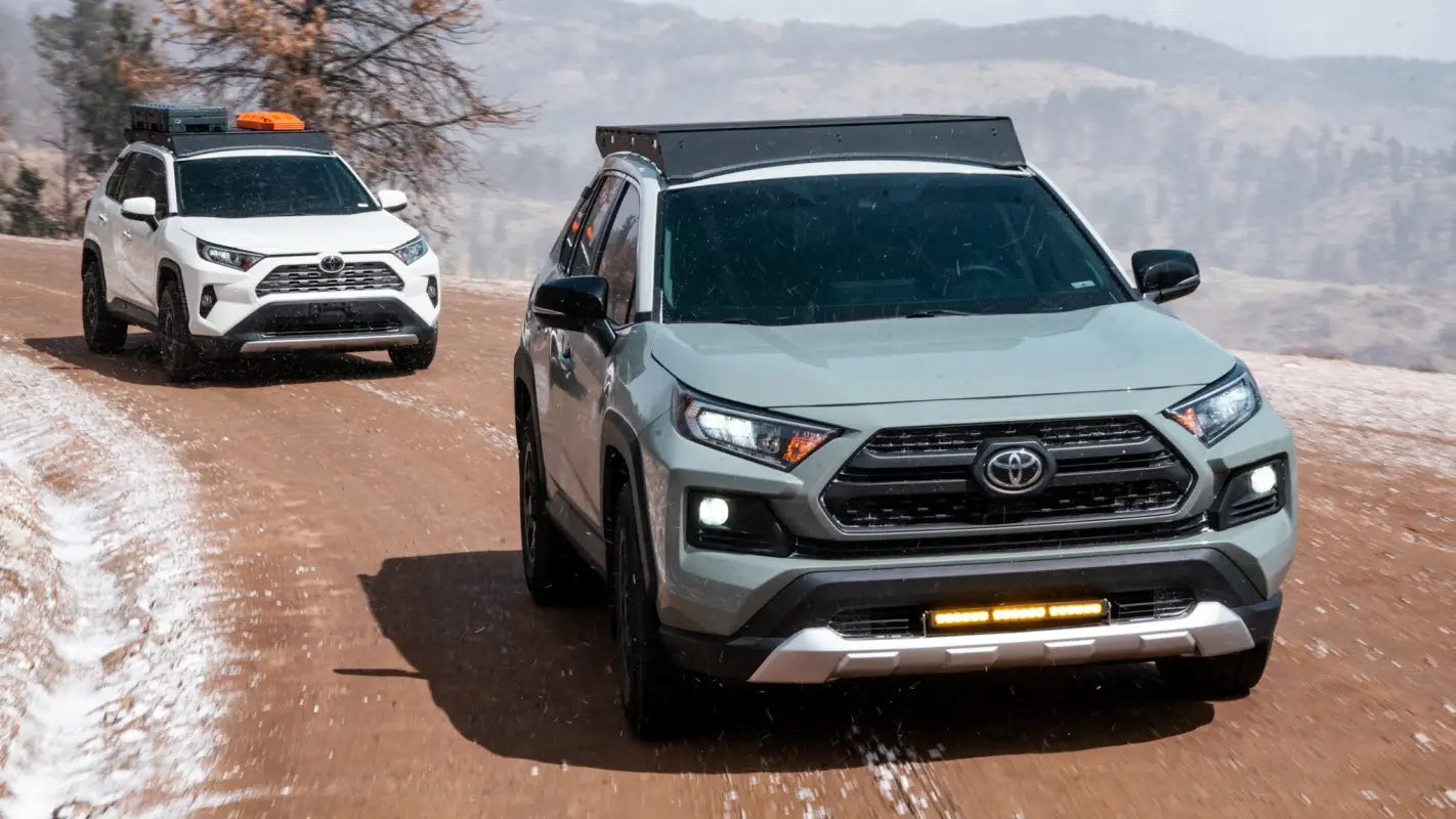 White and gray RAV4s driving on a snowy dirt road, both equipped with The Snowmass roof racks for outdoor adventures.