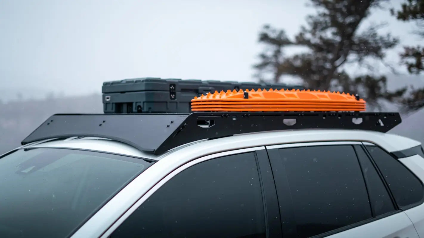 The Snowmass roof rack on a white RAV4, featuring a black aluminum frame with orange recovery boards and a storage box in a snowy setting.