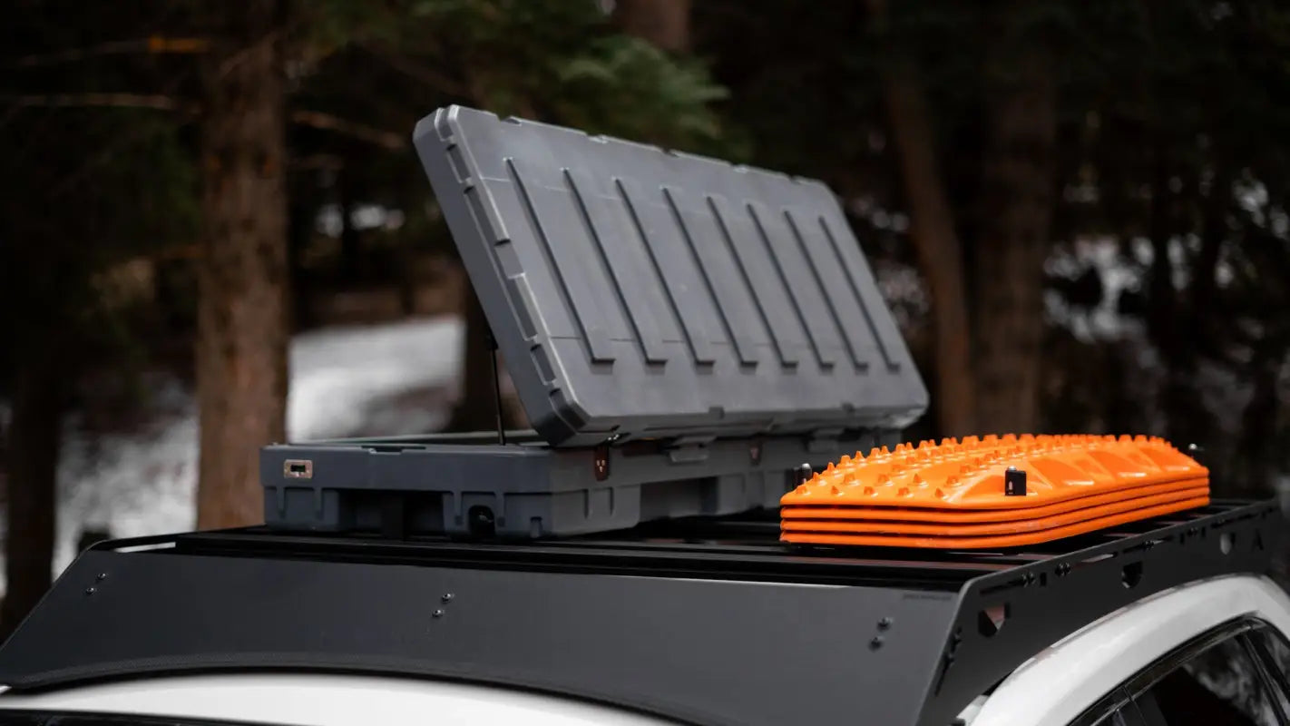 Open storage box and orange recovery boards mounted on The Snowmass roof rack on a white RAV4 in a forest setting.