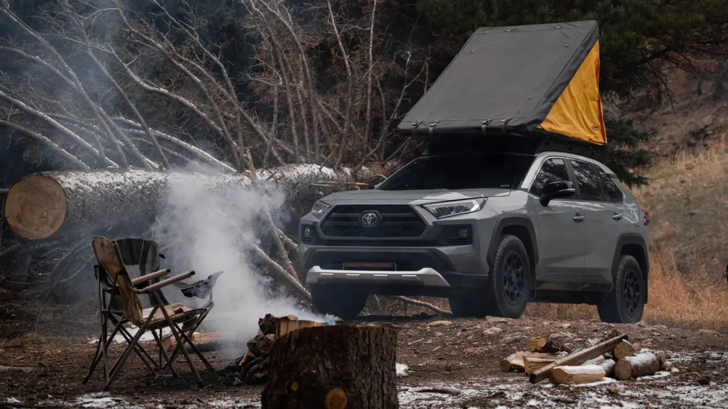 Gray RAV4 with The Snowmass roof rack and rooftop tent set up at a forest campsite, emphasizing outdoor adventure capabilities.