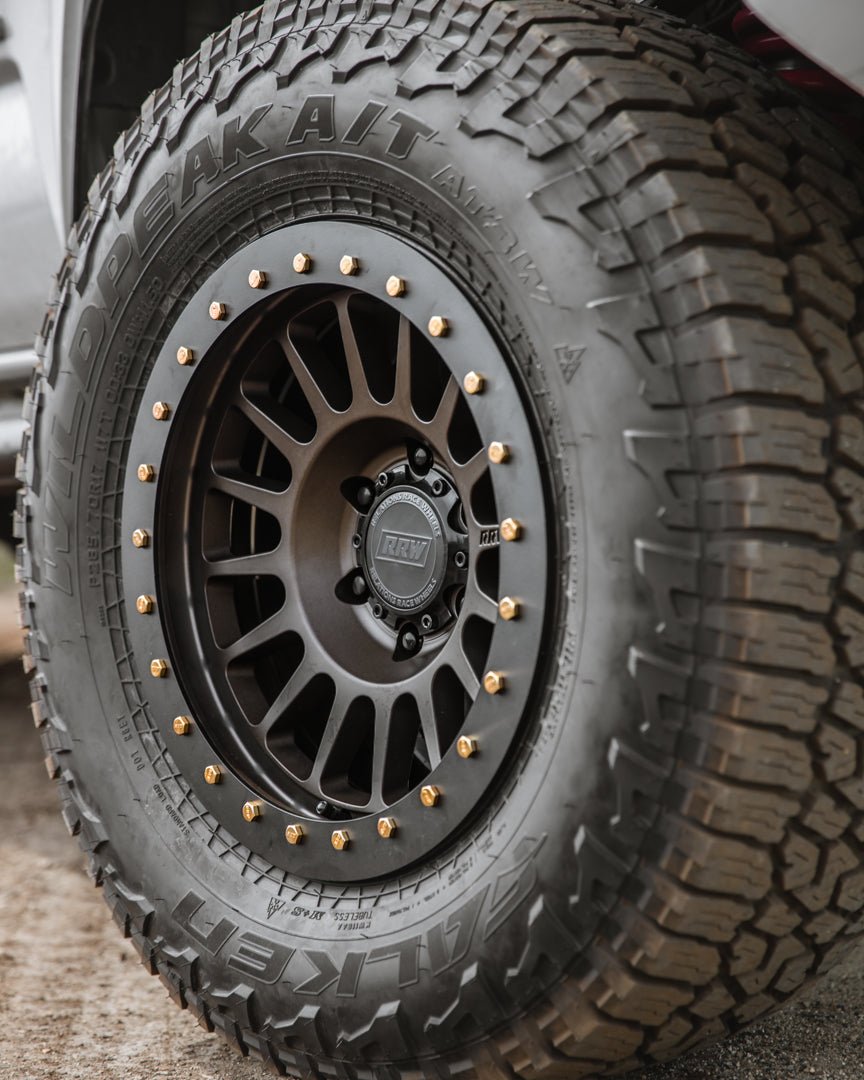 Side view of a hybrid wheel with a black protection ring and gold bolts, installed on an all-terrain tire for rugged use.