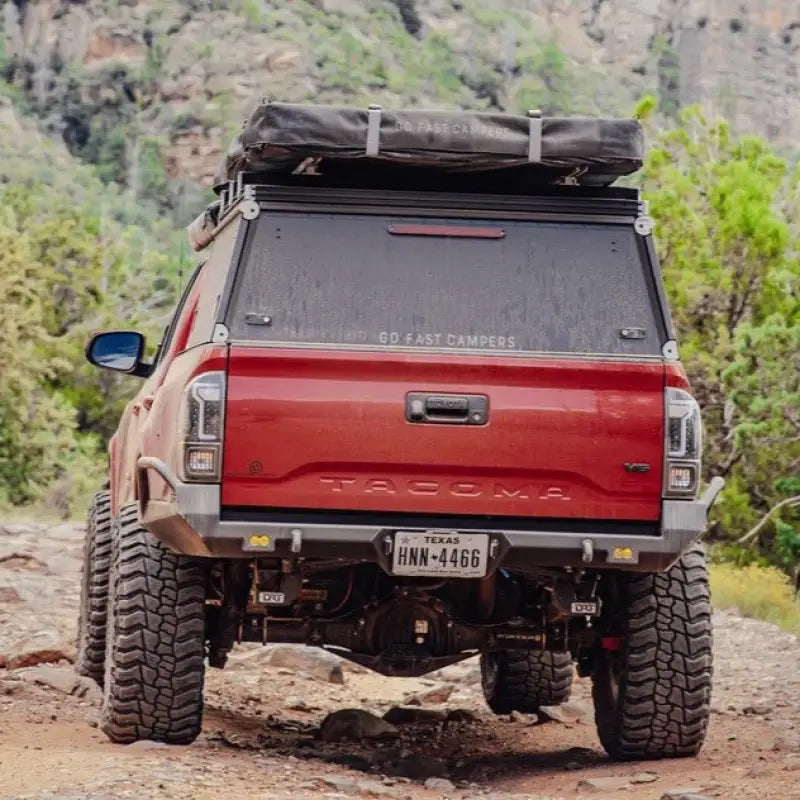 Red Toyota Tacoma with DRT high clearance rear bumper, off-road setup, and rooftop tent in a rugged outdoor setting.