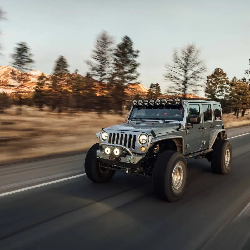 Jeep equipped with Gravity® Titan™ 6" LED lights and roof-mounted light bar, driving on a scenic road at sunset.