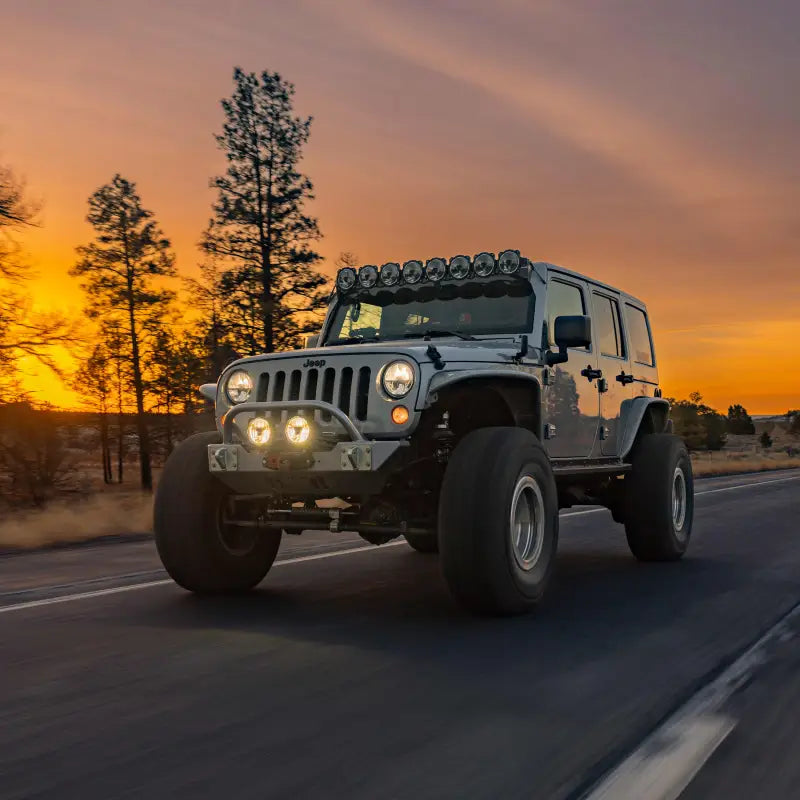 Jeep with Gravity® Titan™ 6" LED lights and roof-mounted light bar, driving on a scenic road during a vibrant sunset.