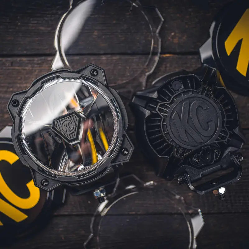 Gravity® Titan™ 6-inch LED off-road lights with KC logo covers, displayed on a wooden surface, highlighting their rugged design.