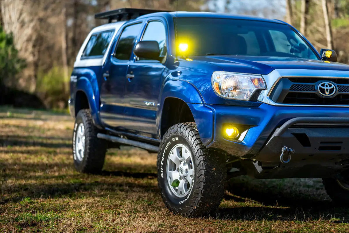 Close-up of blue Toyota Tacoma with Morimoto 4Banger LED Fog Lights in white, illuminating the surroundings in a grassy area.