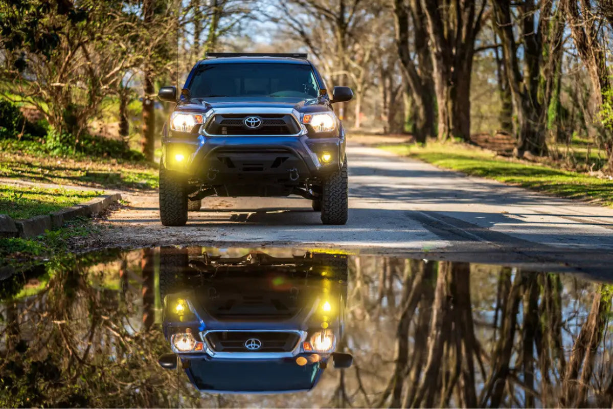 Front view of a blue Toyota Tacoma with Morimoto 4Banger LED Fog Lights, reflecting in a puddle on a tree-lined road.