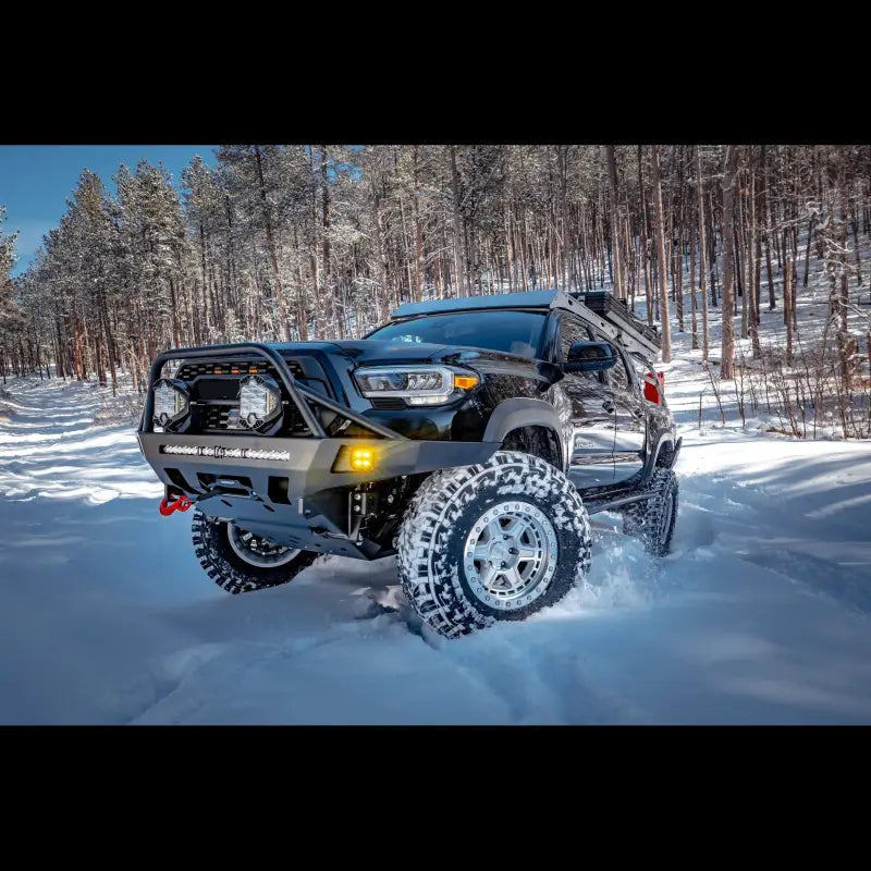 Side-front view of a Toyota Tacoma with Overland Front Bumper, winch, and light bar navigating snow-covered trails. Fits 2016+ models.