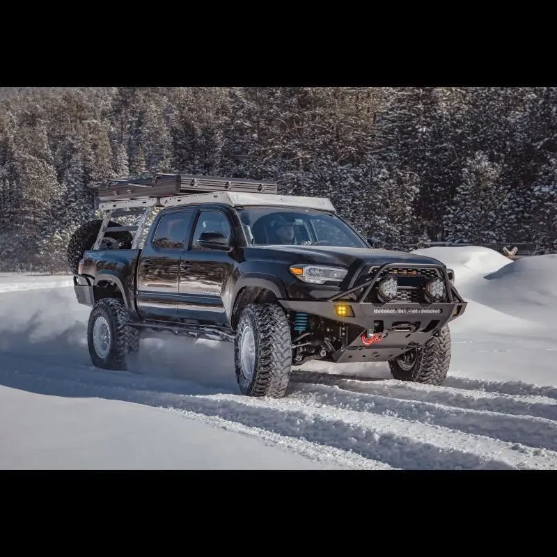 Black Toyota Tacoma with Overland Front Bumper, winch, and light bar driving through snowy terrain. Fits 2016+ models.