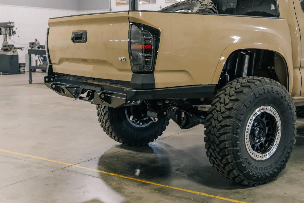 High-clearance Tacoma Rock Runner rear bumper on a beige 3rd Gen Tacoma, displayed in a workshop with off-road tires.