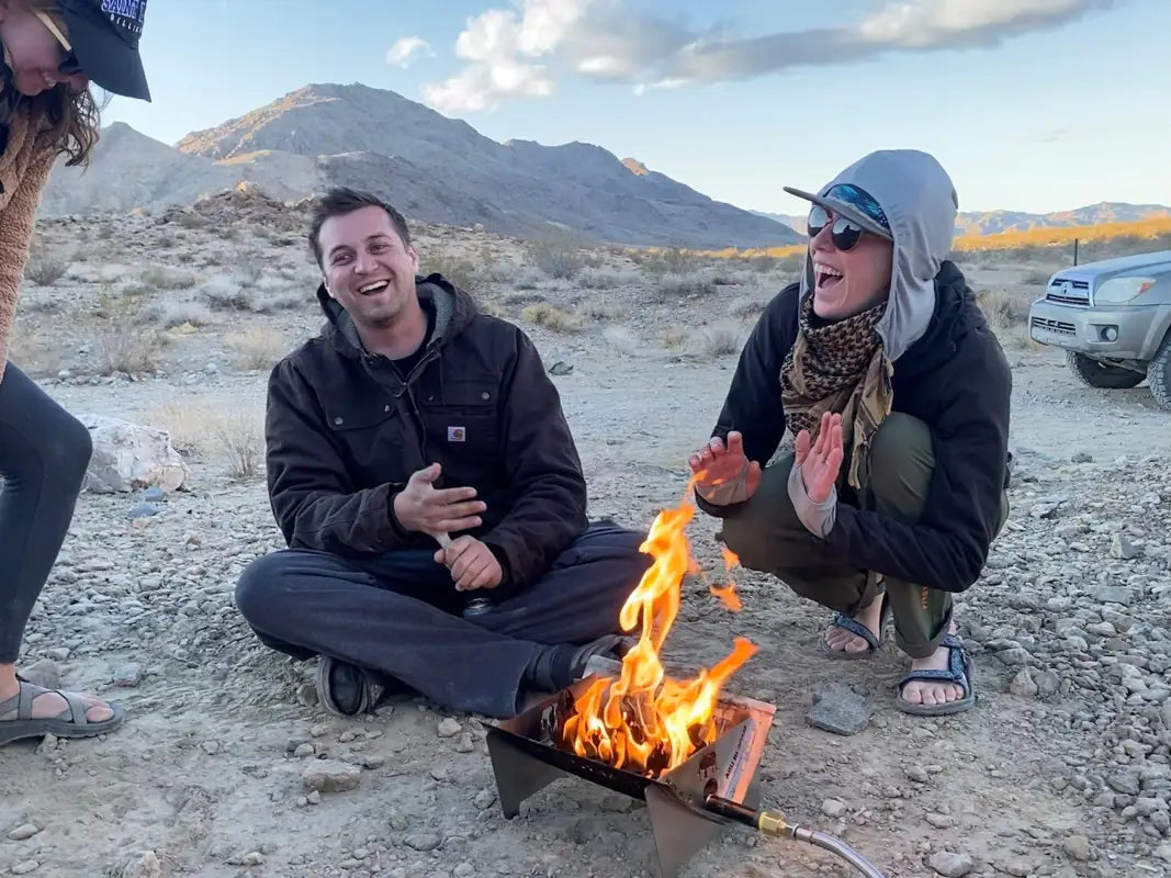 Two people enjoying the TemboTusk Fire Pit outdoors in a desert setting, showcasing its compact and portable propane design for camping.